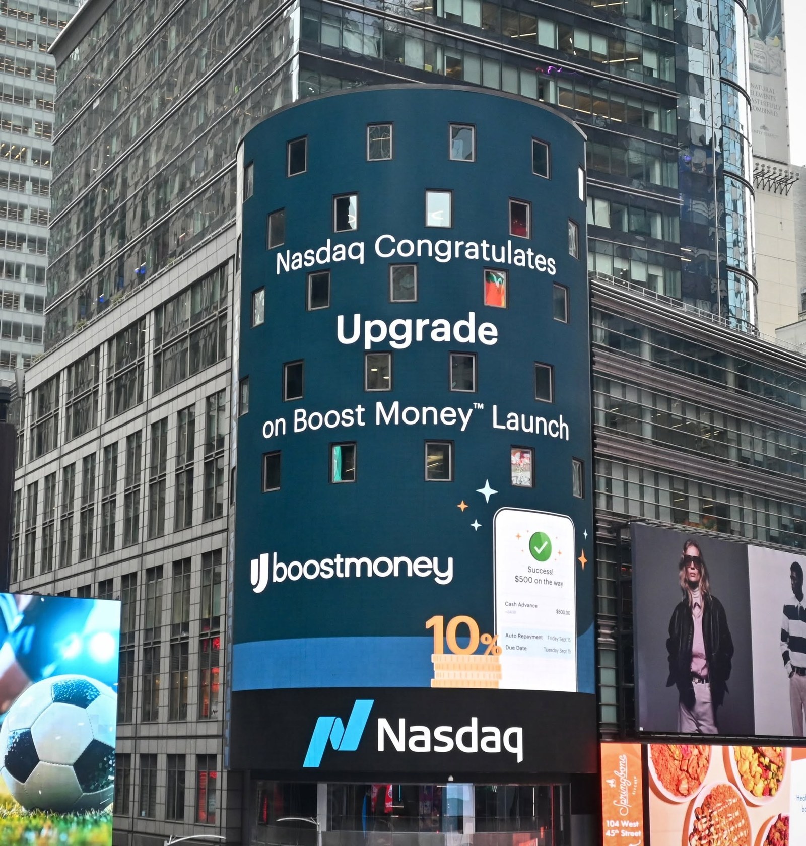 Nasdaq MarketSite tower in Times Square showing the message Nasdaq Congratulates Upgrade on Boost Money Launch, with the Boost Money logo and a phone screen showing a $500 cash advance on the way